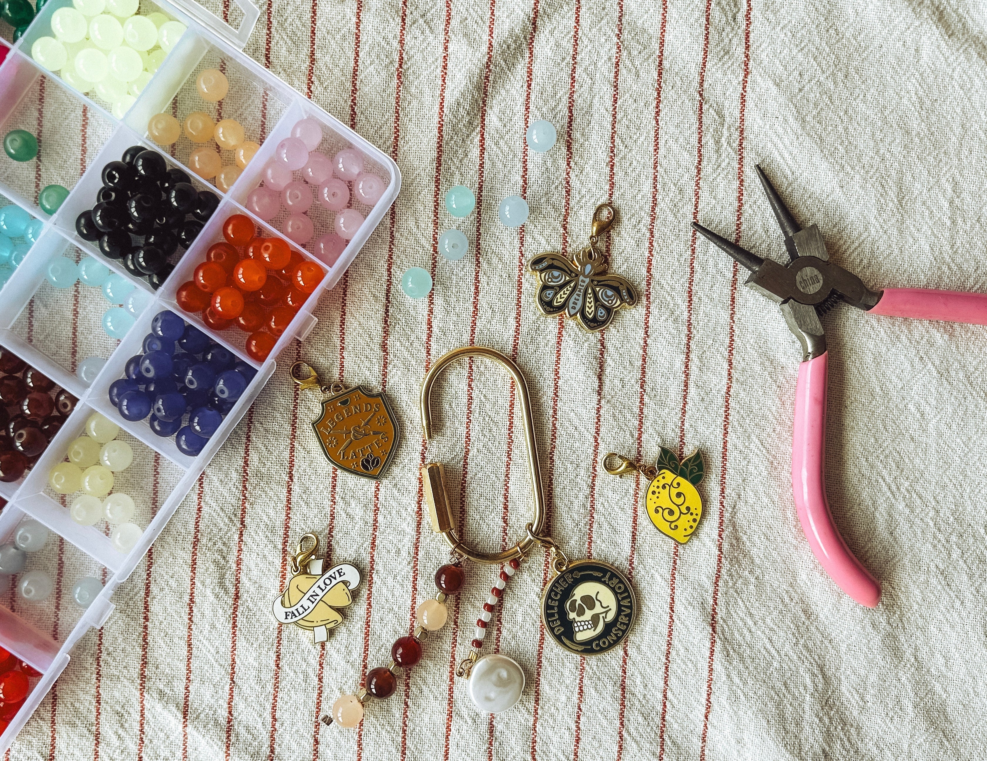 A flat-lay of handmade jewelry charms and beads on a striped fabric background, alongside a plastic bead organizer and pink-handled pliers.